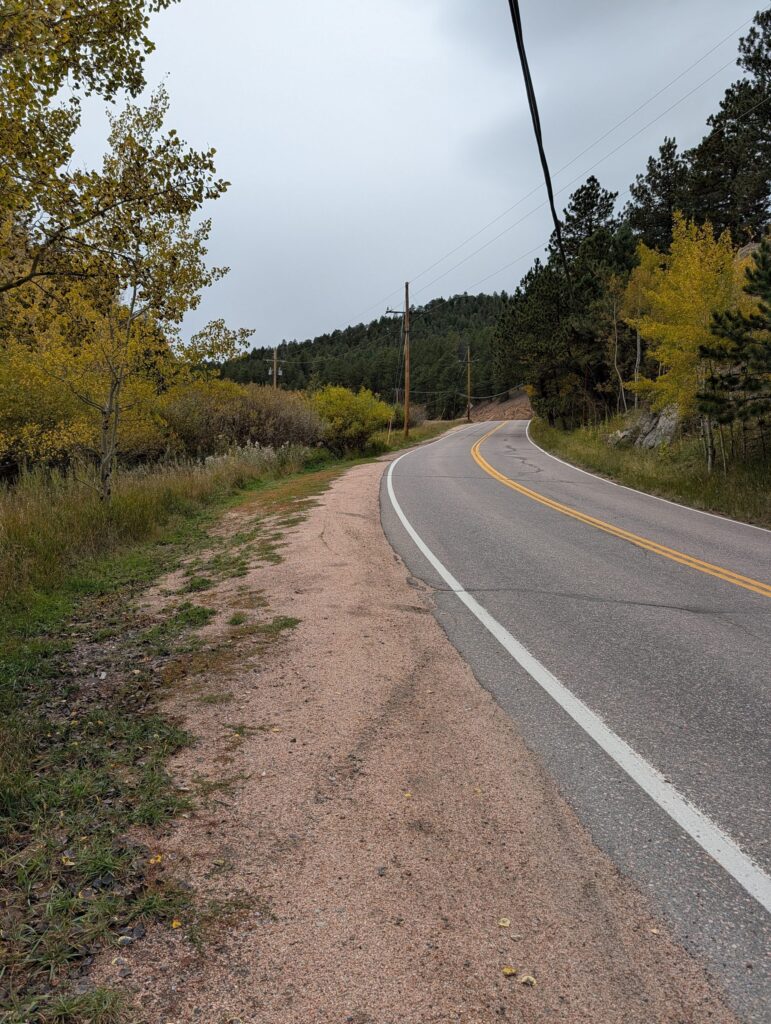 A gravel shoulder along a curving two-lane road with a white edge line, beside aspens turning gold.