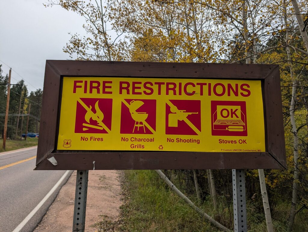 A yellow Forest Service fire restrictions sign beside Brook Forest Road with symbols for no fires, no charcoal grills, no shooting, and stoves OK.