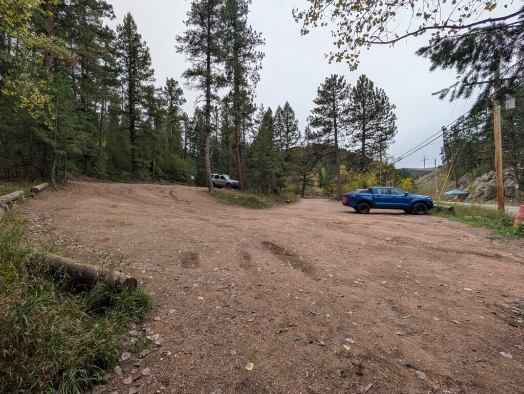The dirt main parking lot at the lower trailhead, mostly empty, edged by pines and log barriers.