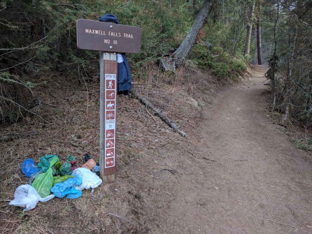 The brown Forest Service post at the lower trailhead reading Maxwell Falls Trail No. 111, with bags of trash and dog waste dumped at its base.