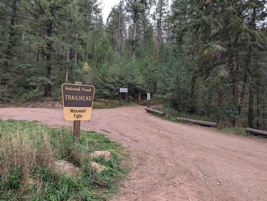 A brown National Forest Trailhead sign for Maxwell Falls at the edge of the dirt parking lot.
