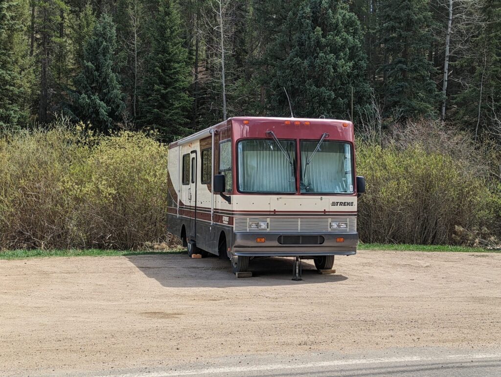 A motorhome set up on leveling blocks with its stabilizer jack down in the gravel overflow lot at the lower trailhead.