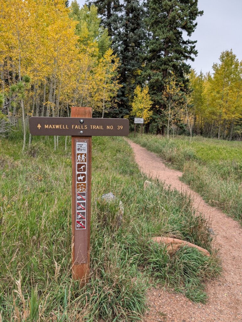 A Forest Service trail post reading Maxwell Falls Trail No. 39 beside a narrow path through grass and aspens.