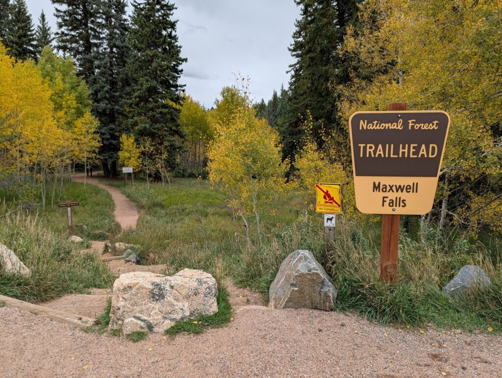 A brown National Forest Trailhead sign for Maxwell Falls beside a path into golden aspens.
