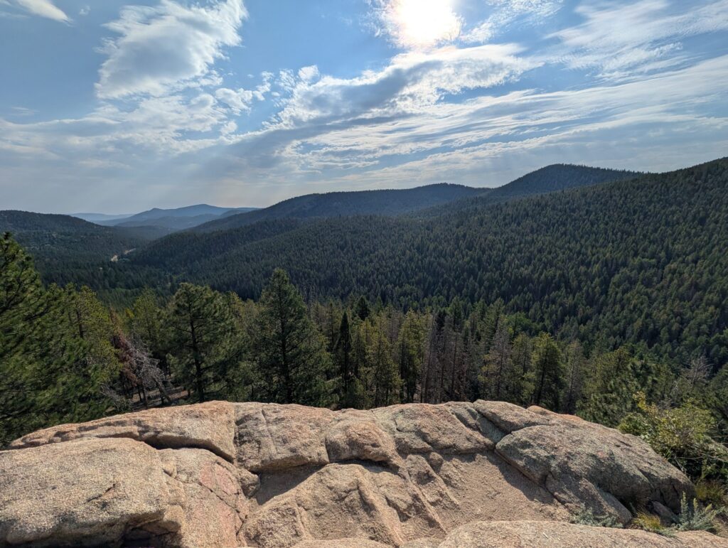 A wide view over forested hills and valleys from a flat granite outcrop under a bright sky.